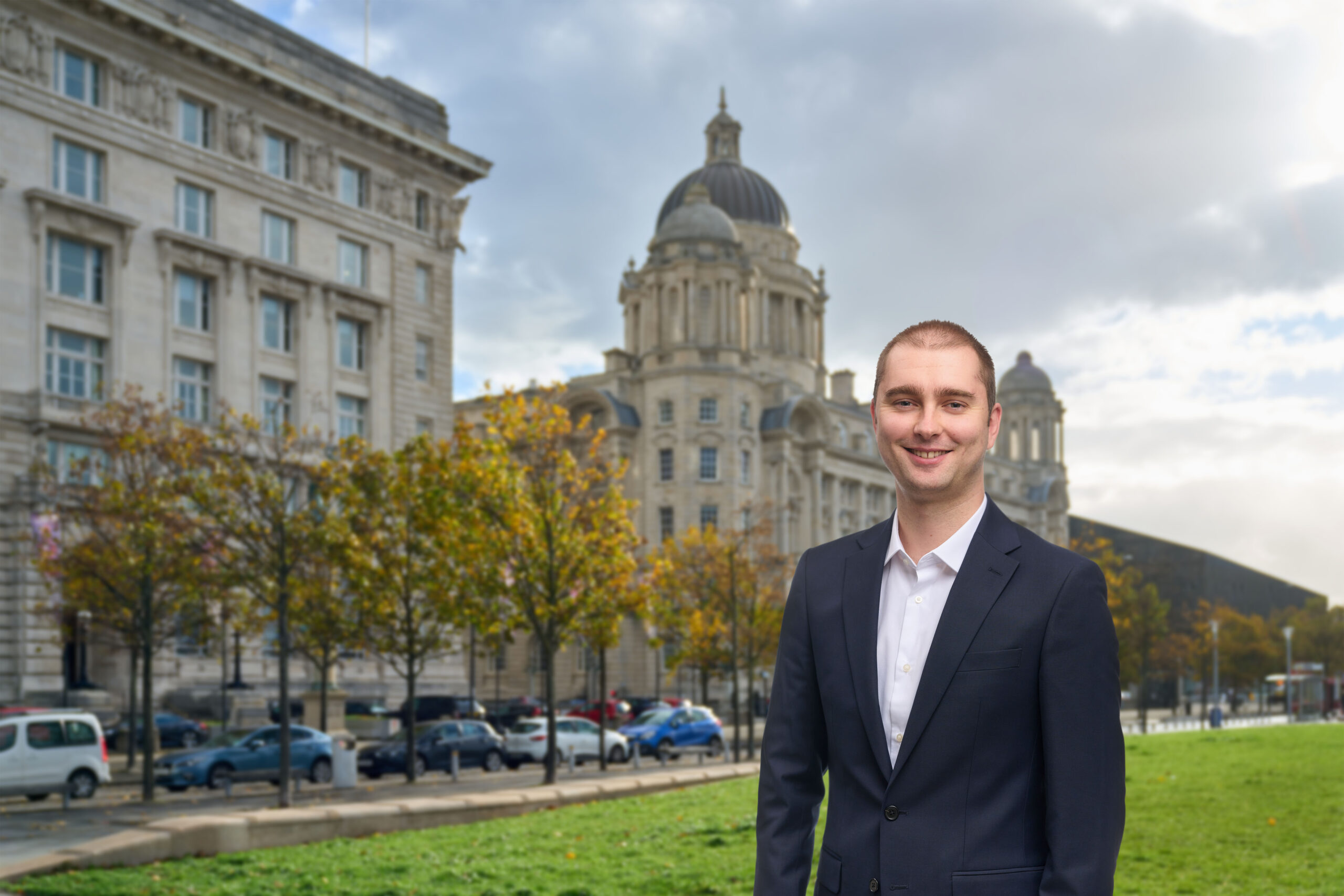 A man in a navy suit jacket and white shirt is standing and smiling in front of the Port of Liverpool building.