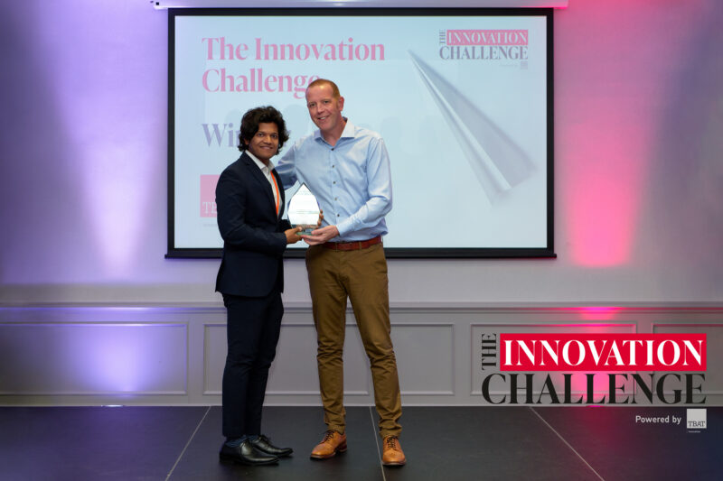 A man in a black suit and white shirt is being handed a glass trophy by a man wearing a blue shirt and light brown trousers. They are both smiling and standing on a small stage in front of a screen which reads "The Innovation Challenge - Winner".