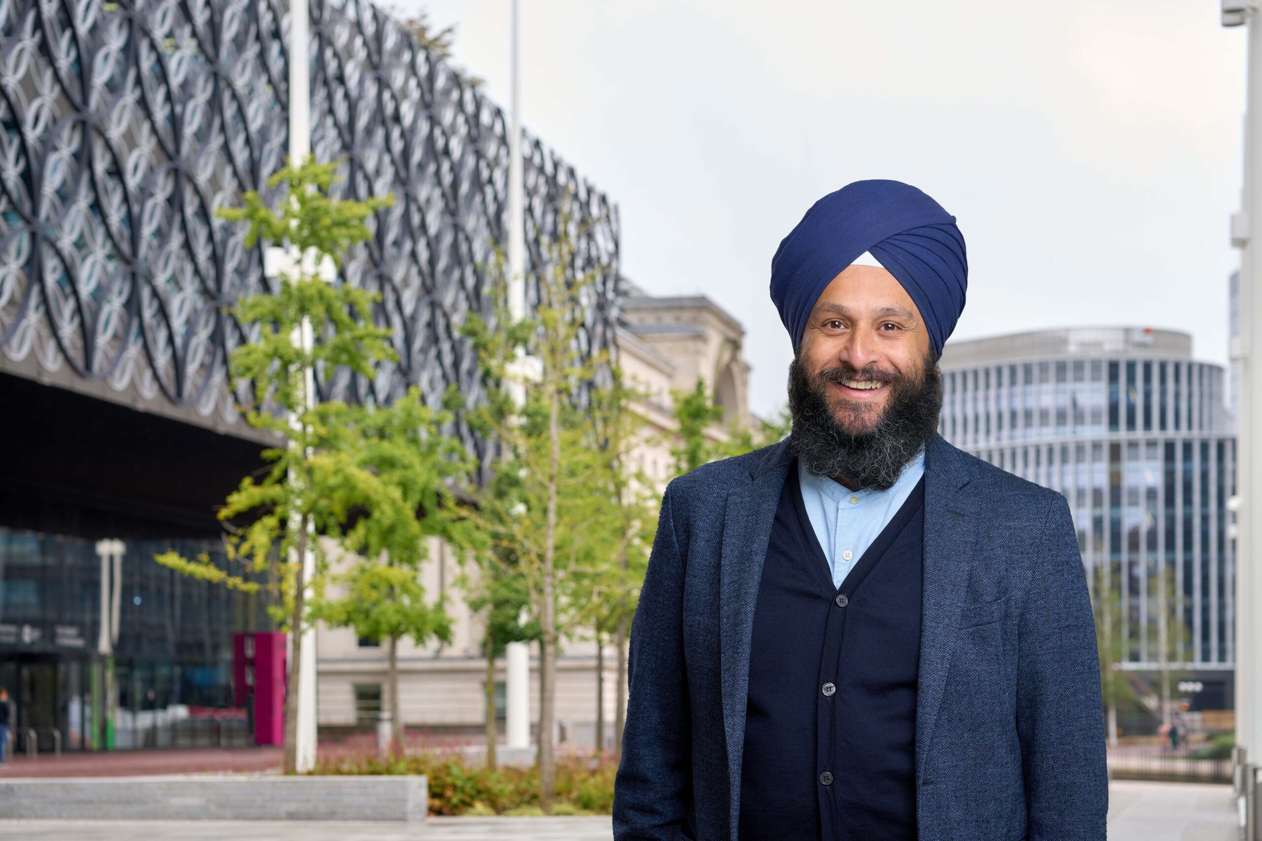 A man in a blue shirt, navy cardigan and blue suit jacket with a blue turban and beard is smiling and standing in front of some trees and buildings in central Birmingham.