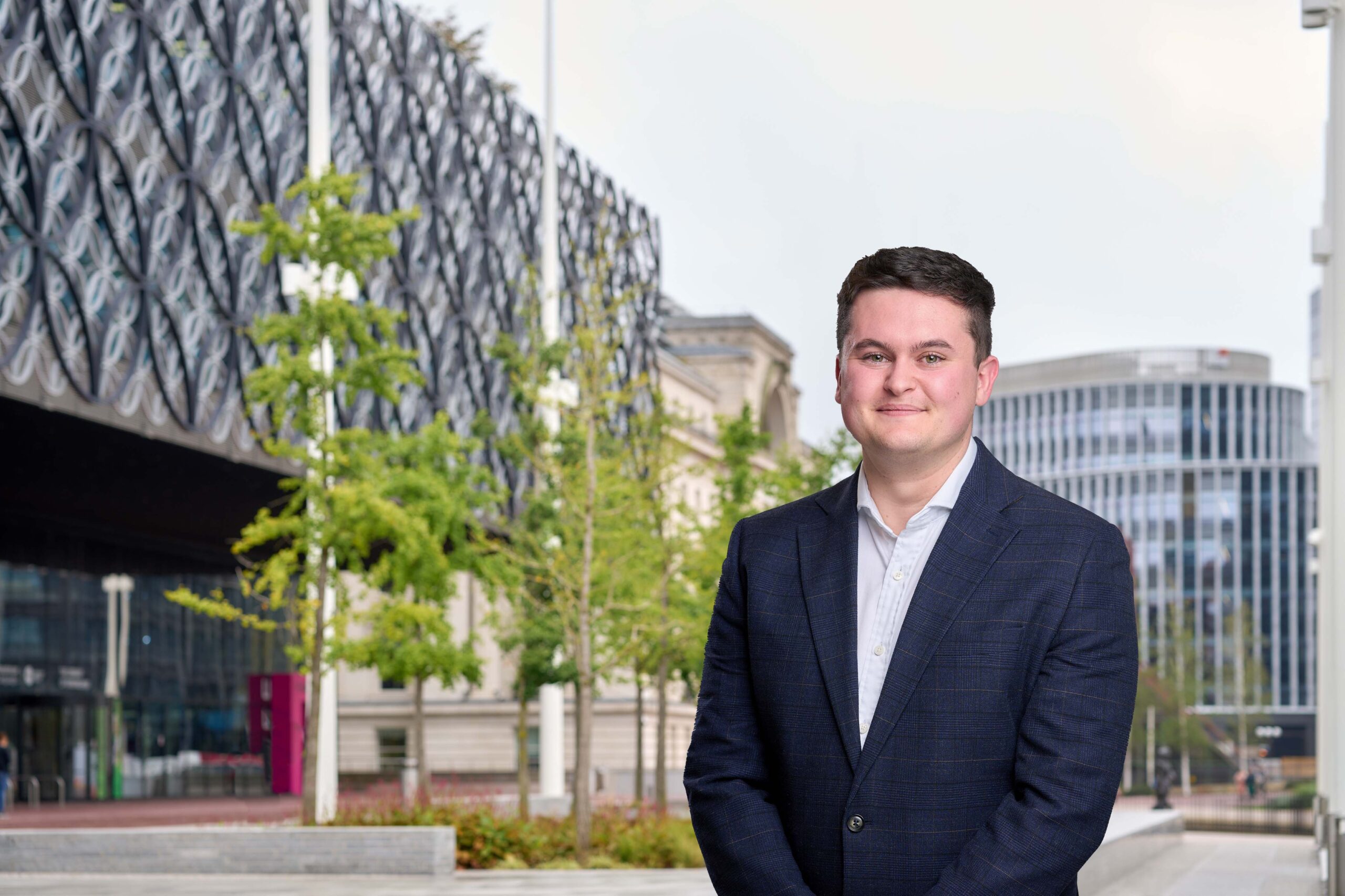 A man in a white shirt and black suit jacket is standing and smiling in front of some trees and buildings in central Birmingham.